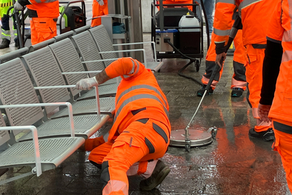 Mehrere Personen reinigen eine Haltestelle. Sie nutzen unterschiedliche Mittel und Geräte zum Putzen: einen Lappen, einen Hochdruckreiniger und ein spezielles Reinigungsgerät mit Düse. Die Personen tragen orangefarbene Warnkleidung. 