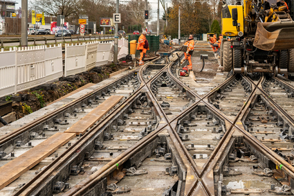 Gleisbauarbeiten an einer Straßenbahnkreuzung mit mehreren spitz zulaufenden Schienenprofilen im Zentrum, an denen Personen in Warnkleidung arbeiten. Ein gelbes Baustellenfahrzeug steht seitlich an den Gleisen.
