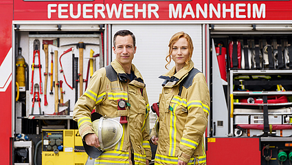 Eine Feuerwehrfrau und ein Feuerwehrmann vor einem Feuerwehrwagen.