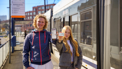 Zwei Personen stehen an einer Straßenbahnhaltestelle neben einer haltenden Rhein-Neckar-Tram (RNT). Sie halten ein Smartphone mit einer geöffneten Ticket‑App und eine Deutschland-Ticket-Chipkarte in die Kamera.