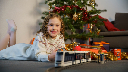 Ein Mädchen mit dunkelblonden Korkenzieher-Locken in einem Strickkleid liegt vor einem festlich geschmückten Weihnachtsbaum und lächelt. Im Vordergrund fährt das Klemmbaustein-Modell einer Straßenbahn vom Typ Rhein-Neckar-Tram (RNT).