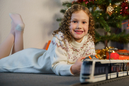 Ein Mädchen mit dunkelblonden Korkenzieher-Locken in einem Strickkleid liegt vor einem festlich geschmückten Weihnachtsbaum und lächelt. Im Vordergrund fährt das Klemmbaustein-Modell einer Straßenbahn vom Typ Rhein-Neckar-Tram (RNT).