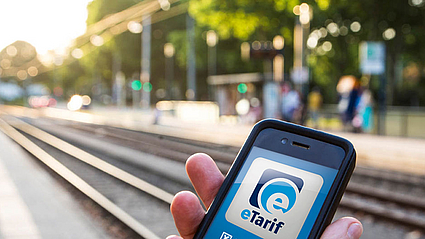 Hand hält Smartphone an einer Stadtbahnhaltestelle, auf dem Handy ist das Logo der eTarif-App zu sehen