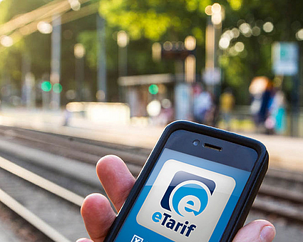 Hand hält Smartphone an einer Stadtbahnhaltestelle, auf dem Handy ist das Logo der eTarif-App zu sehen