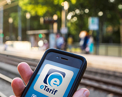 Hand hält Smartphone an einer Stadtbahnhaltestelle, auf dem Handy ist das Logo der eTarif-App zu sehen