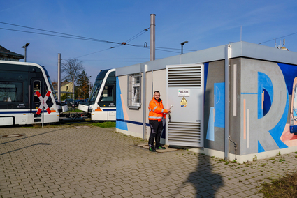 Ein Mitarbeiter der rnv in oranger Sicherheitskleidung steht in der geöffneten Tür eines Gleichrichterunterwerks und blick in die Kamera. Im Hintergrund fährt eine Straßenbahn vom Typ Rhein-Neckar-Tram vorbei.