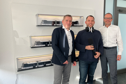 Die Geschäftsführer der Rhein-Neckar-Verkehr GmbH (rnv), Martin in der Beek und Christian Volz stehen links und rechts neben Moderator Jens Schneider in einem Besprechungszimmer. An der Wand hinter ihnen sind drei Modelle der Rhein-Neckar-Tram in den drei Längenausführungen (30, 40 und 60 Meter) auf Regalbrettchen ausgestellt.