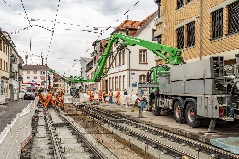 Auf der Dossenheimer Landstraße werden zwei neue Gleise betoniert. Ein Spezialfahrzeug transportiert Beton ins Gleisbett. Zahlreiche Gleisarbeiter sind beteiligt.