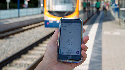 Handy an einer Haltestelle mit Stadtbahn im Hintergrund 