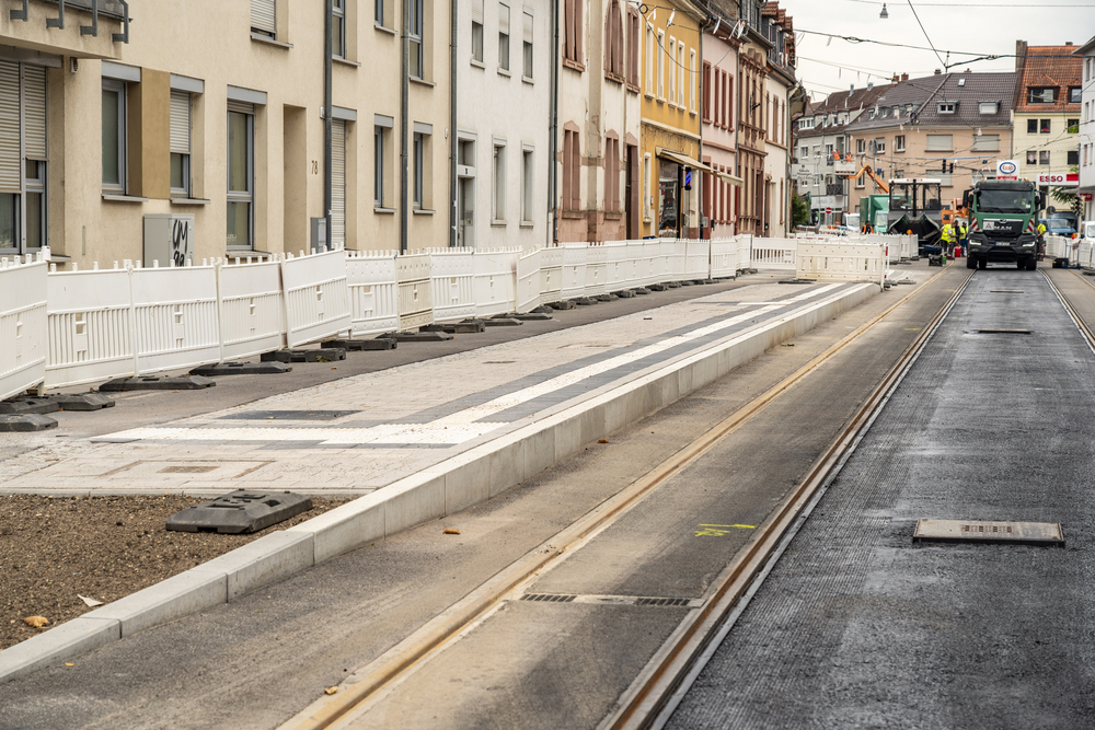 In der Dossenheimer Landstraße sind frisch asphaltierte Gleise und der noch mit Barken abgesperrte neue barrierefreie Steig der Haltestelle Johann-Fischer-Straße zu sehen. Im Hintergrund stehen Baufahrzeuge.
